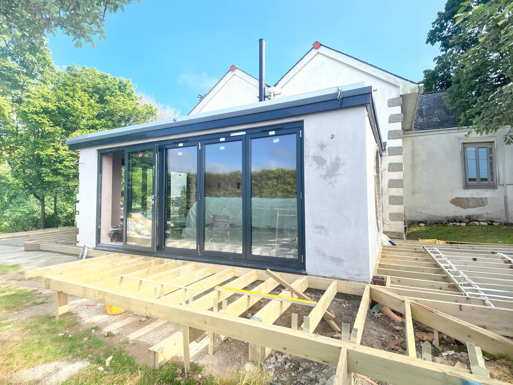 A single-storey extension under construction, with anthracite aluminium bi-fold doors installed, fresh white render meeting the original granite quoins, and the timber joists of a new deck being laid in front of it.