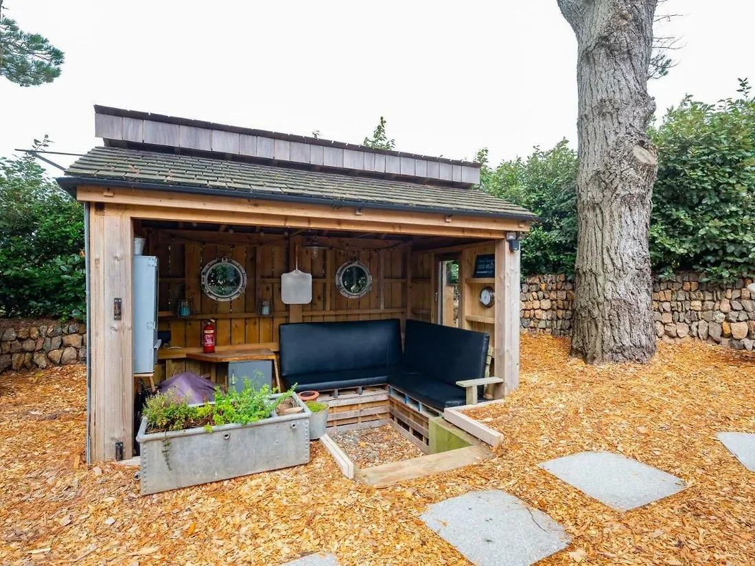 A timber garden bar and shelter with a cedar shingle roof, porthole windows and bench seating, set against a granite wall.